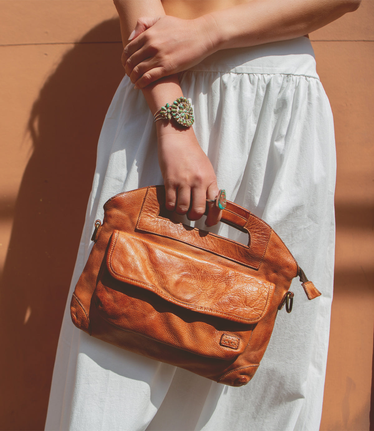 A person in a white skirt holds the Bed|Stü Greenway brown full-grain leather handbag with cutout handles and a zip-top closure, accessorizing with a large green ring and matching bracelet while standing against a peach-colored background.