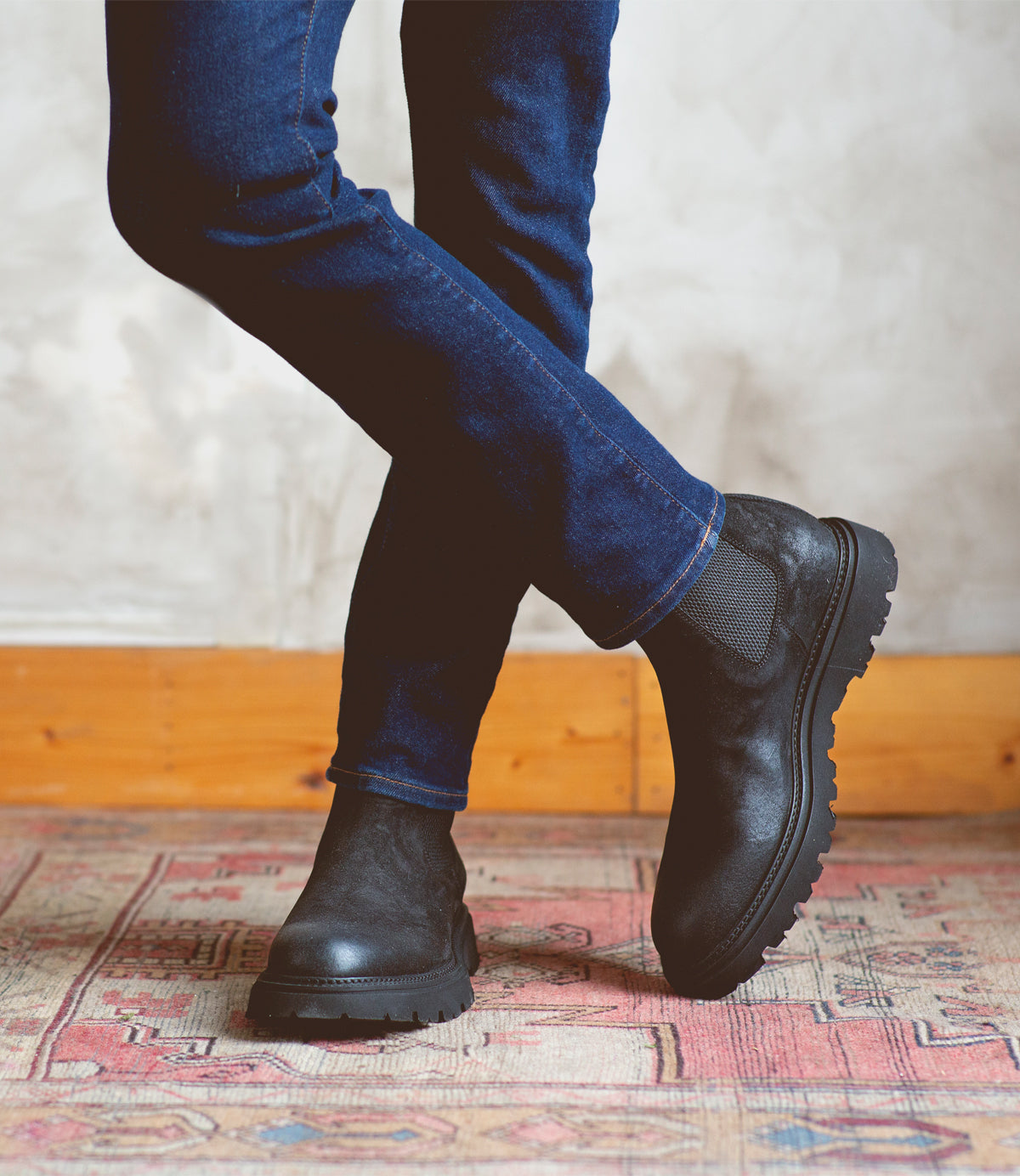A person wearing dark jeans and Bed|Stü’s Grit black men’s Chelsea boots stands with legs crossed on a patterned rug, against a light-colored wall and wooden baseboard.