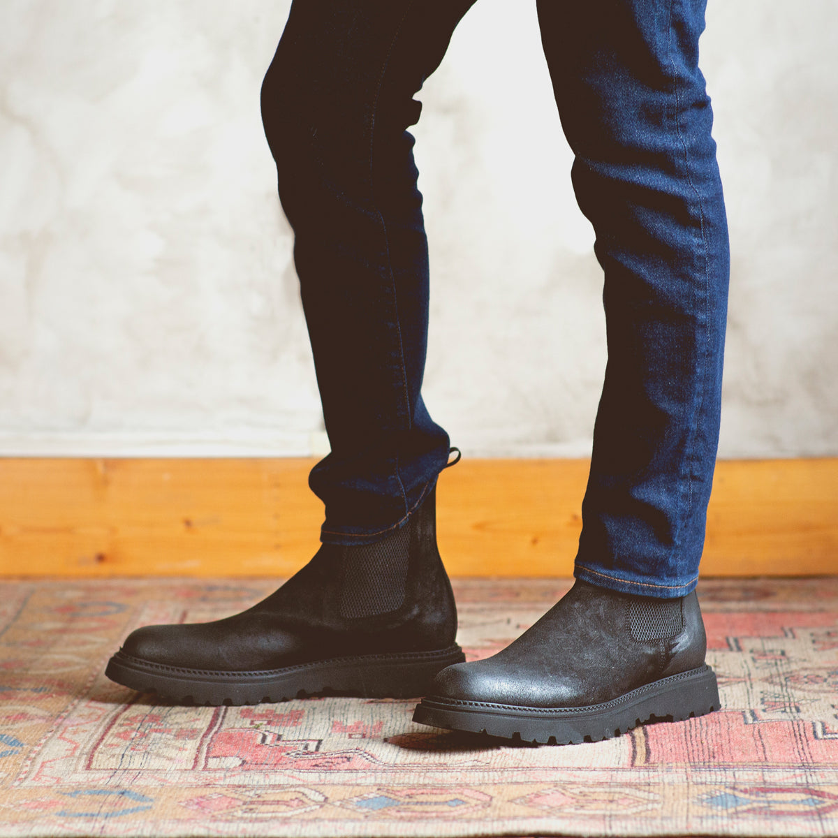A person wearing dark blue jeans and black Bed|Stü Grit Chelsea boots stands on a patterned rug next to a light-colored wall with wooden baseboard. These Italian handcrafted Grit boots by Bed|Stü add a refined touch to the understated ensemble.