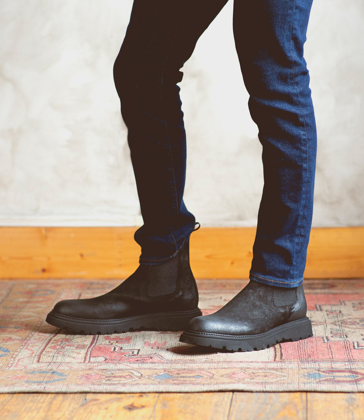 A person wearing dark blue jeans and black Bed|Stü Grit Chelsea boots stands on a patterned rug next to a light-colored wall with wooden baseboard. These Italian handcrafted Grit boots by Bed|Stü add a refined touch to the understated ensemble.