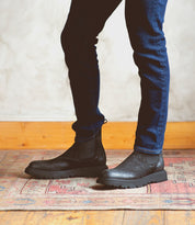 A person wearing dark blue jeans and black Bed|Stü Grit Chelsea boots stands on a patterned rug next to a light-colored wall with wooden baseboard. These Italian handcrafted Grit boots by Bed|Stü add a refined touch to the understated ensemble.