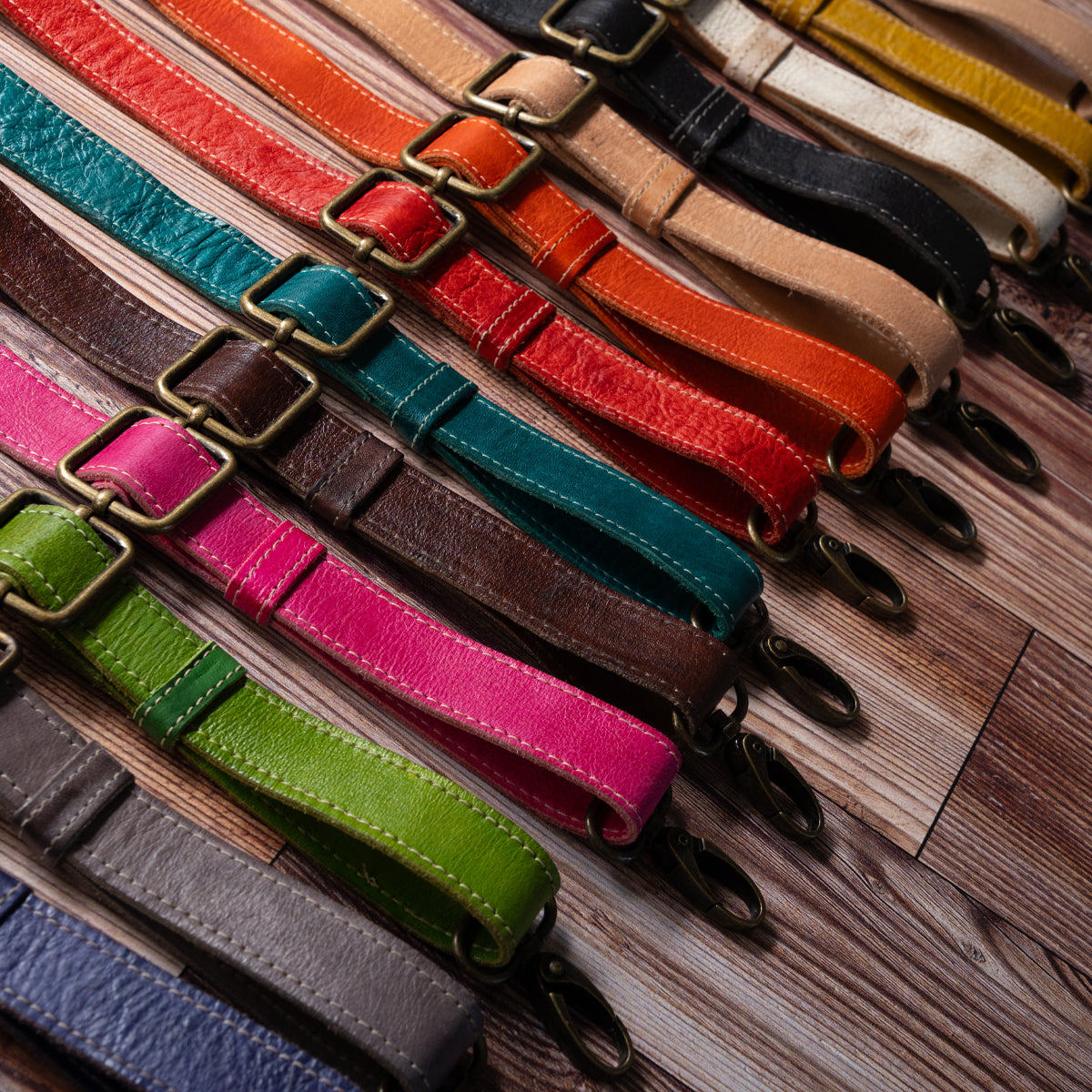 A selection of colorful Bed|Stü Crossbody Straps, each featuring adjustable leather designs with sturdy metal clasps, is displayed diagonally on a wooden surface—ideal for customizing your Bed|Stü bag or adding a fresh crossbody style to your favorite accessory.