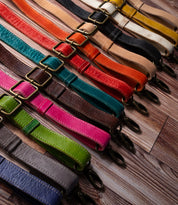 A selection of colorful Bed|Stü Crossbody Straps, each featuring adjustable leather designs with sturdy metal clasps, is displayed diagonally on a wooden surface—ideal for customizing your Bed|Stü bag or adding a fresh crossbody style to your favorite accessory.