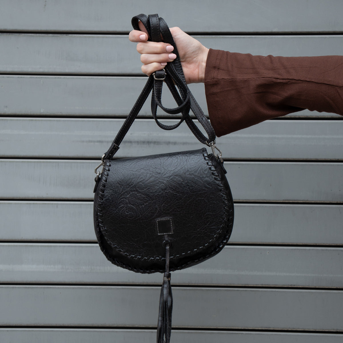 A person wearing a brown long-sleeve top holds up the Bed|Stü Half Moon EM, a black leather saddlebag crossbody purse with a braided edge and tassel, featuring an adjustable strap, against a gray metal background.