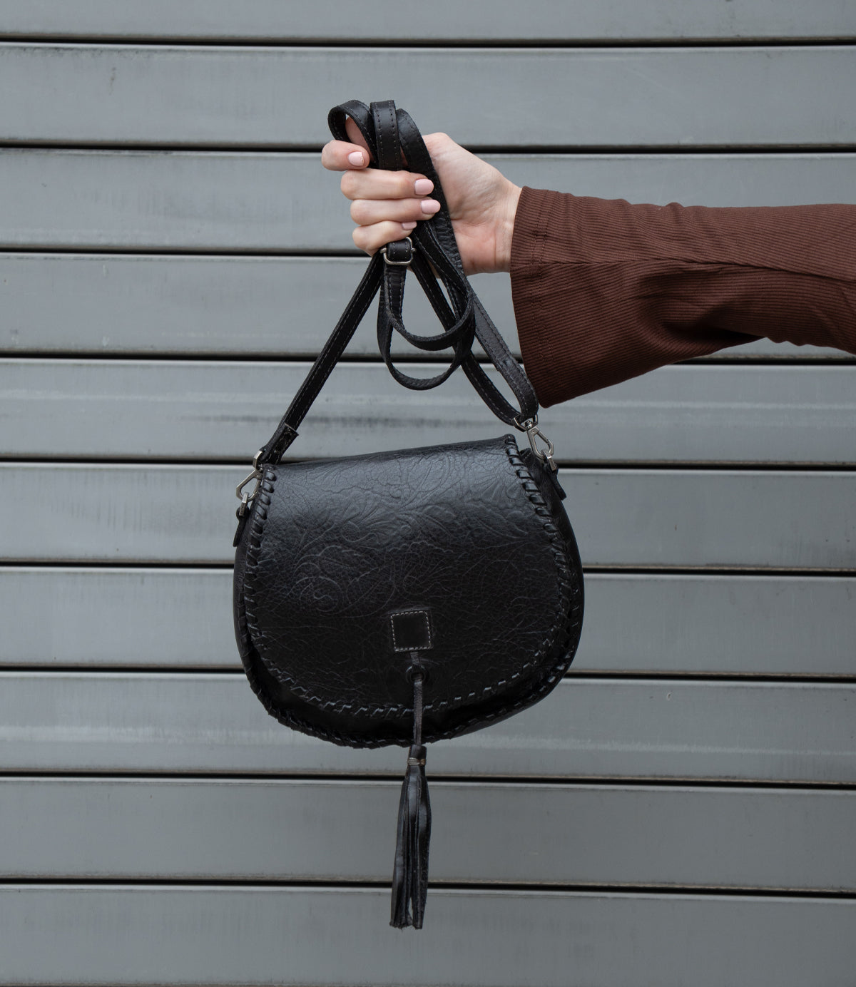 A person wearing a brown long-sleeve top holds up the Bed|Stü Half Moon EM, a black leather saddlebag crossbody purse with a braided edge and tassel, featuring an adjustable strap, against a gray metal background.