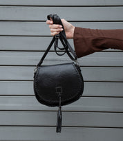 A person wearing a brown long-sleeve top holds up the Bed|Stü Half Moon EM, a black leather saddlebag crossbody purse with a braided edge and tassel, featuring an adjustable strap, against a gray metal background.