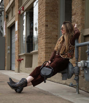 A woman dressed in brown clothing, boots, and sunglasses sits on a metal gas meter against a brick building, holding the Bed|Stü Half Moon EM black saddlebag crossbody purse with an adjustable strap and looks to the side.