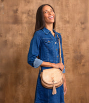 A woman in a blue denim dress stands against a wooden background, smiling and wearing the Bed|Stü Half Moon EM saddlebag crossbody purse, which features an embossed foliage pattern, adjustable strap, and tassel.