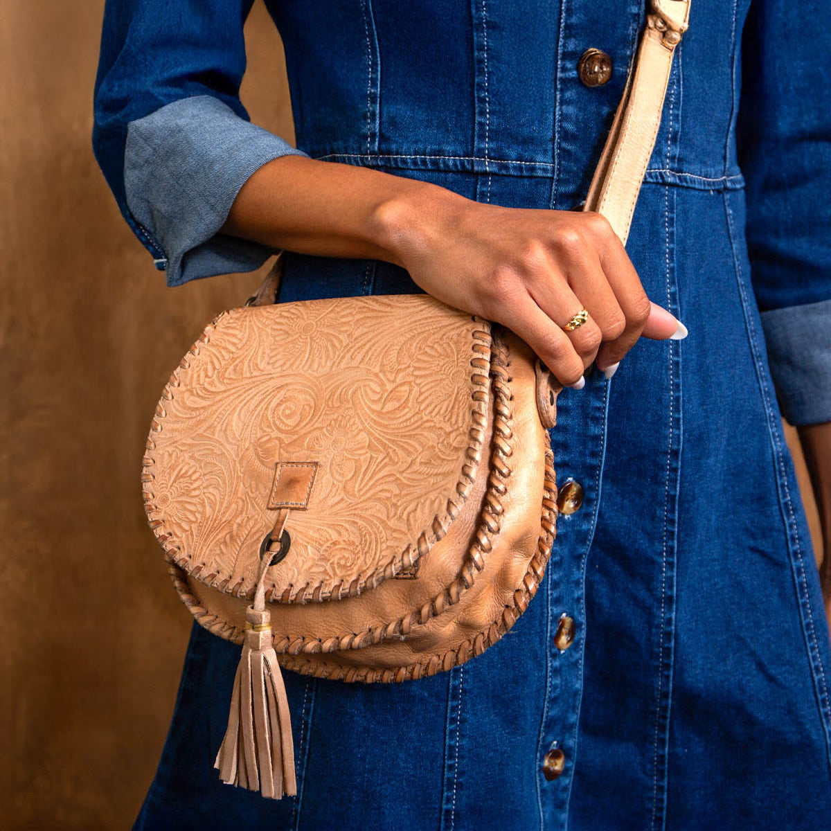 A person wearing a blue denim dress holds the Bed|Stü Half Moon EM, a tan leather saddlebag crossbody purse with an embossed foliage pattern, adjustable strap, and tassel.