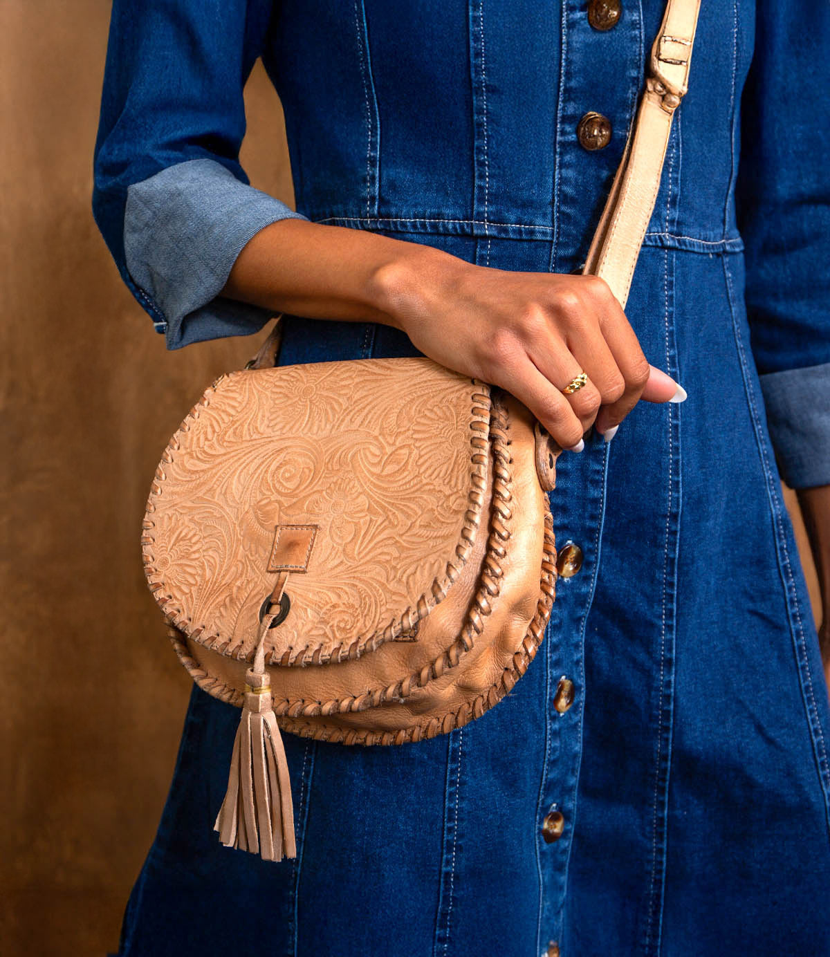 A person wearing a blue denim dress holds the Bed|Stü Half Moon EM, a tan leather saddlebag crossbody purse with an embossed foliage pattern, adjustable strap, and tassel.
