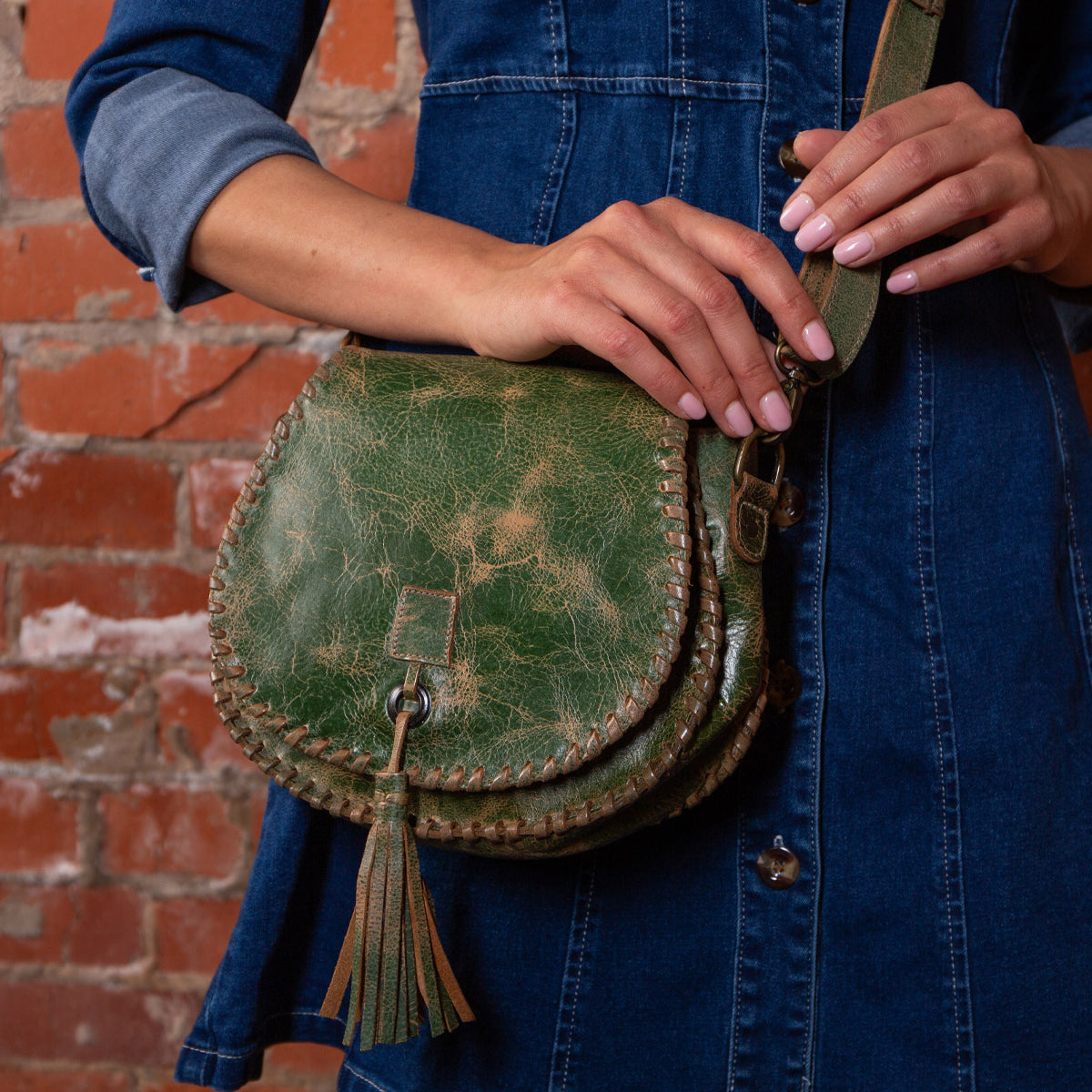 A person in a blue denim dress holds the Bed|Stü Half Moon green leather tassel bag, featuring an adjustable strap and hand-laced stitching, while standing in front of a brick wall.