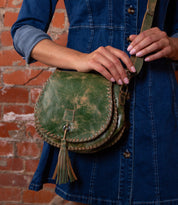 A person in a blue denim dress holds the Bed|Stü Half Moon green leather tassel bag, featuring an adjustable strap and hand-laced stitching, while standing in front of a brick wall.