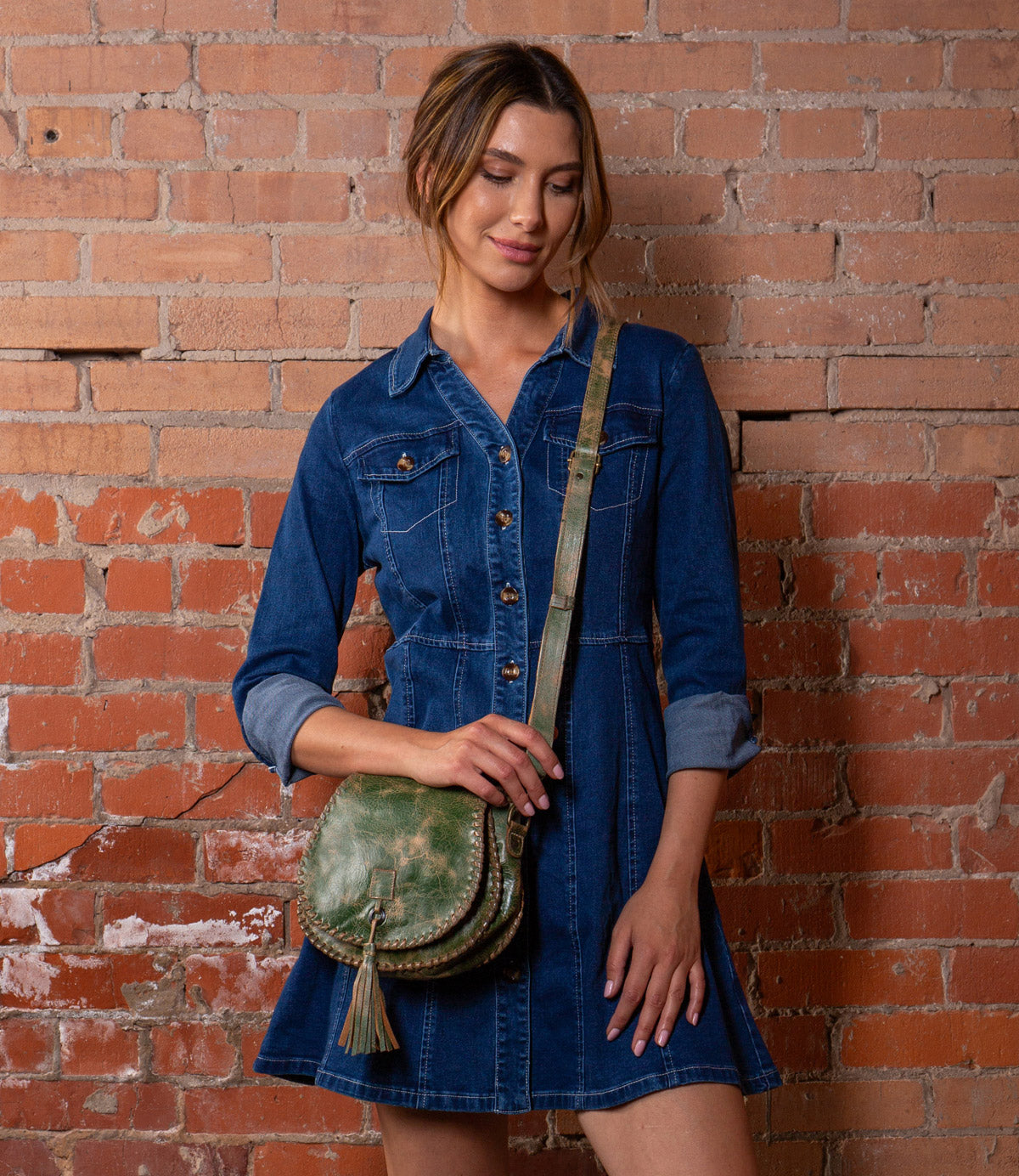 A woman in a denim dress stands against a brick wall, holding the Bed|Stü Half Moon—a green leather tassel bag featuring an adjustable strap and hand-laced stitching.