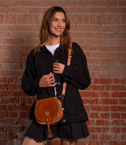 A woman stands against a brick wall, smiling and looking to the side, wearing a black denim jacket, black shorts, and carrying the Bed|Stü Half Moon brown leather tassel bag with an adjustable strap.