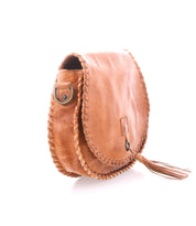The Bed|Stü Half Moon is a tan leather crossbody bag featuring hand-laced whipstitch edges, a metal ring detail on the side, and a tassel accent hanging from the bottom.