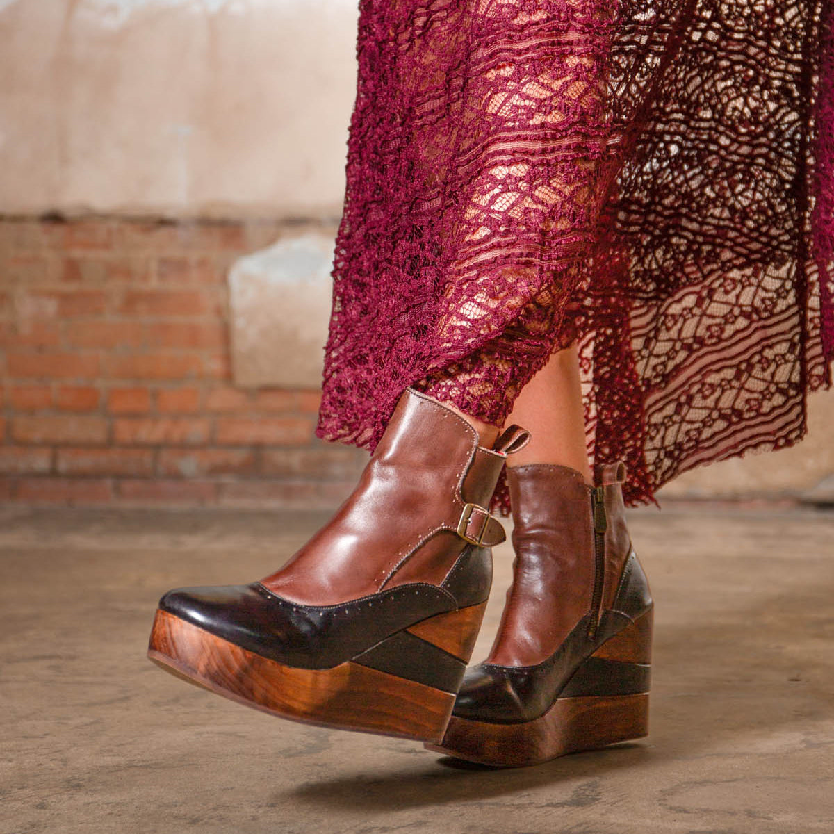 A person is wearing the Bed|Stü Harmonious brown leather platform wedge ankle boots, featuring sculpted wood bases and smooth leather uppers, paired with a burgundy lace skirt and standing on a concrete floor against a brick wall backdrop.