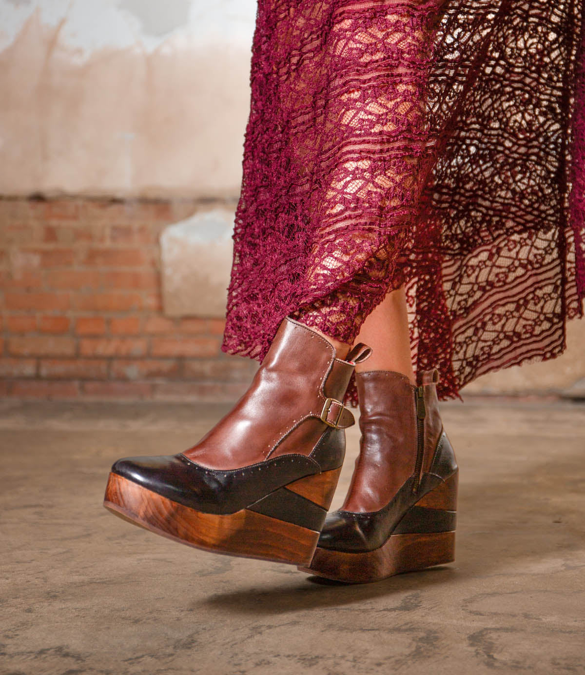 A person is wearing the Bed|Stü Harmonious brown leather platform wedge ankle boots, featuring sculpted wood bases and smooth leather uppers, paired with a burgundy lace skirt and standing on a concrete floor against a brick wall backdrop.