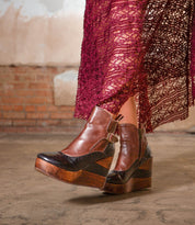 A person is wearing the Bed|Stü Harmonious brown leather platform wedge ankle boots, featuring sculpted wood bases and smooth leather uppers, paired with a burgundy lace skirt and standing on a concrete floor against a brick wall backdrop.