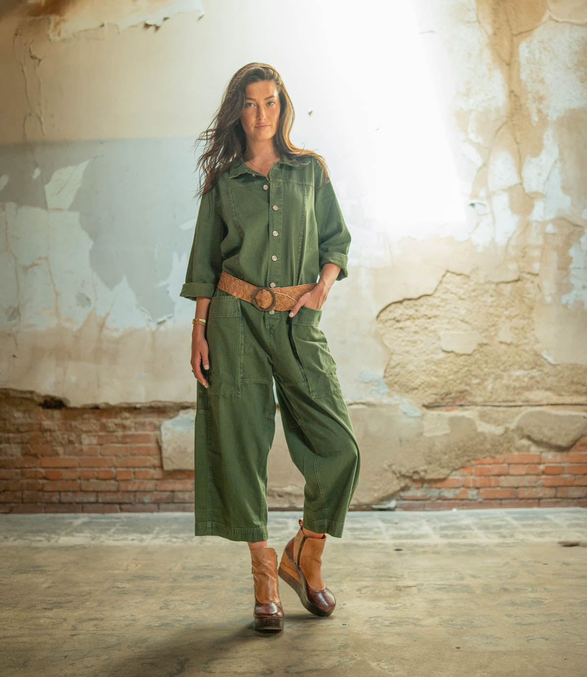 A woman stands indoors wearing a green jumpsuit, cinched at the waist with a brown belt and complemented by Bed|Stü's Harmonious platform wedge ankle boots featuring sculpted wood bases. The background highlights a peeling, textured wall and brickwork at the base.