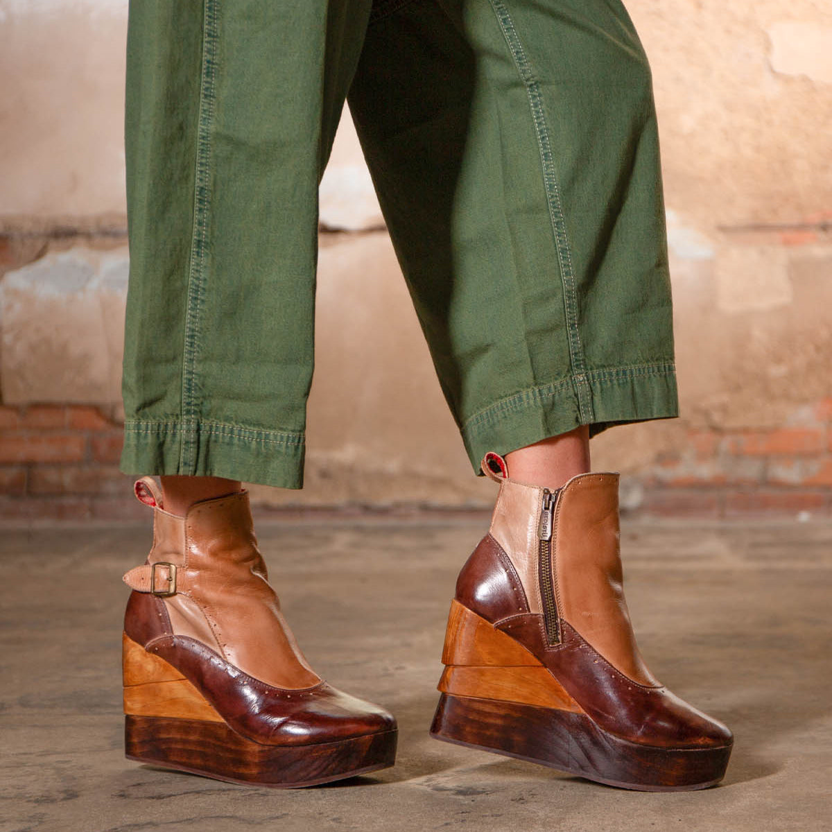 A person wearing green pants and the Bed|Stü Harmonious stylish platform wedge ankle boots—featuring sculpted wood bases, leather uppers, a zipper, and buckle—stands on a concrete floor in front of a rustic brick wall.