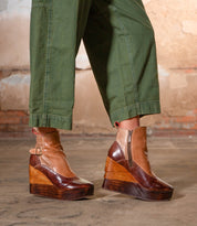 A person wearing green pants and the Bed|Stü Harmonious stylish platform wedge ankle boots—featuring sculpted wood bases, leather uppers, a zipper, and buckle—stands on a concrete floor in front of a rustic brick wall.