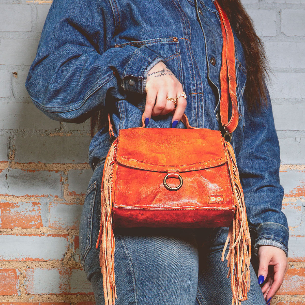 A person in a denim jacket and jeans holds the Bed|Stü Hidden—an orange leather fringed handbag featuring a structured silhouette and crossbody strap—while standing against a light brick wall.