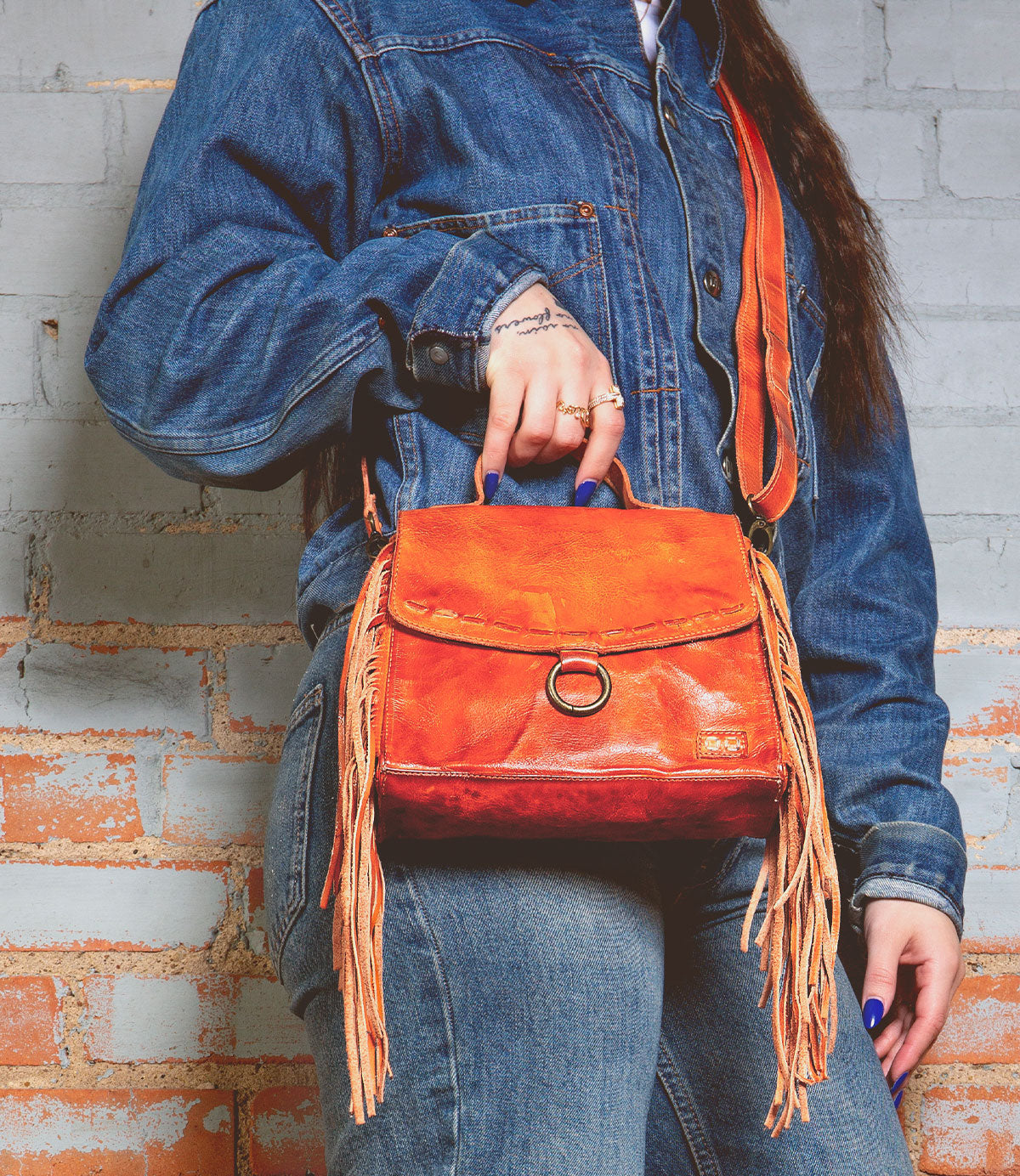 A person in a denim jacket and jeans holds the Bed|Stü Hidden—an orange leather fringed handbag featuring a structured silhouette and crossbody strap—while standing against a light brick wall.
