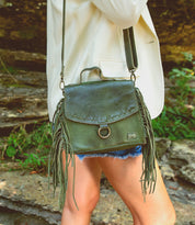 A person in a white blazer and denim shorts carries the Bed|Stü Hidden green leather fringe crossbody bag with a structured silhouette outdoors near rocks and greenery.
