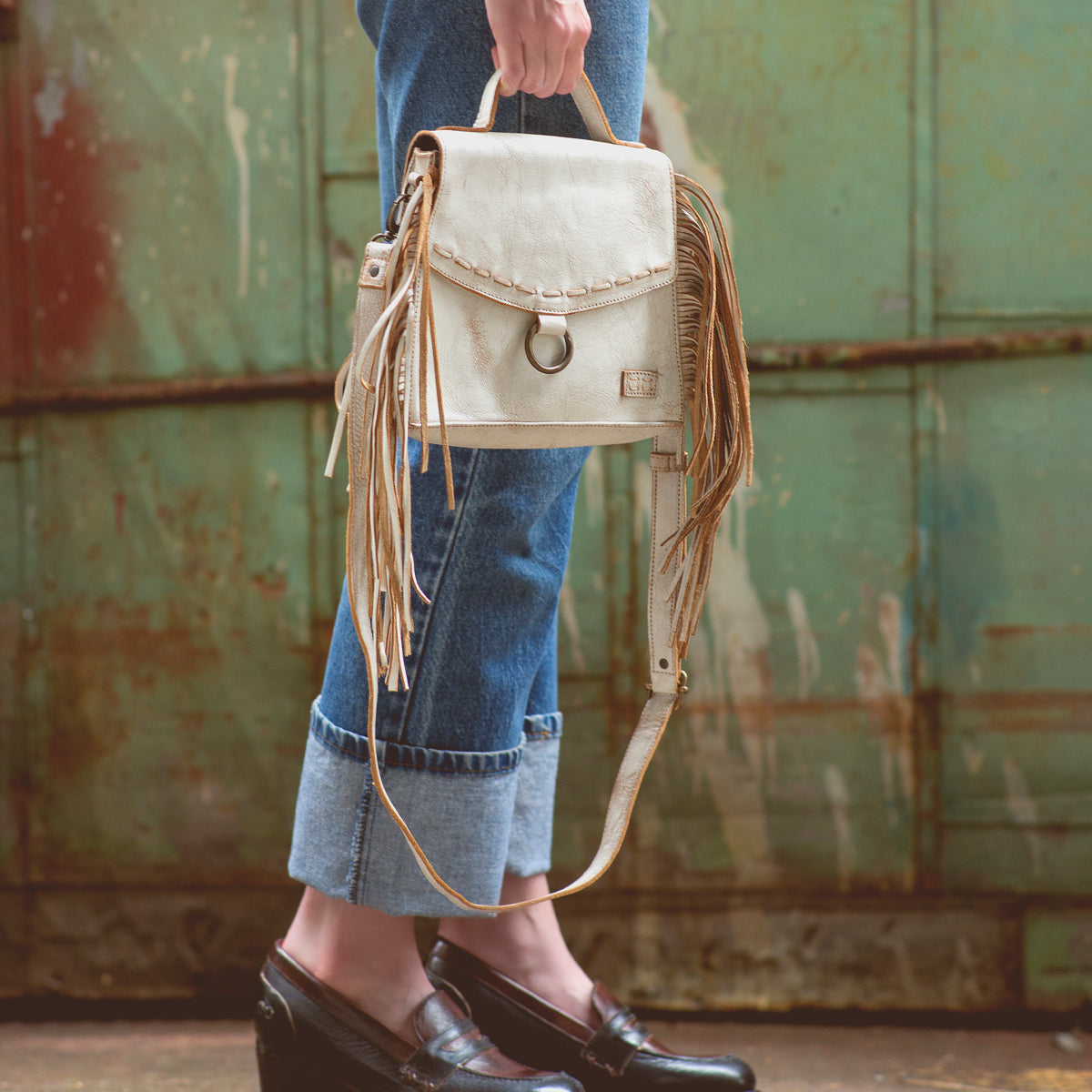 A person wearing cuffed jeans and black loafers holds the Bed|Stü Hidden beige fringed handbag with a structured silhouette in front of a weathered, green metal background.