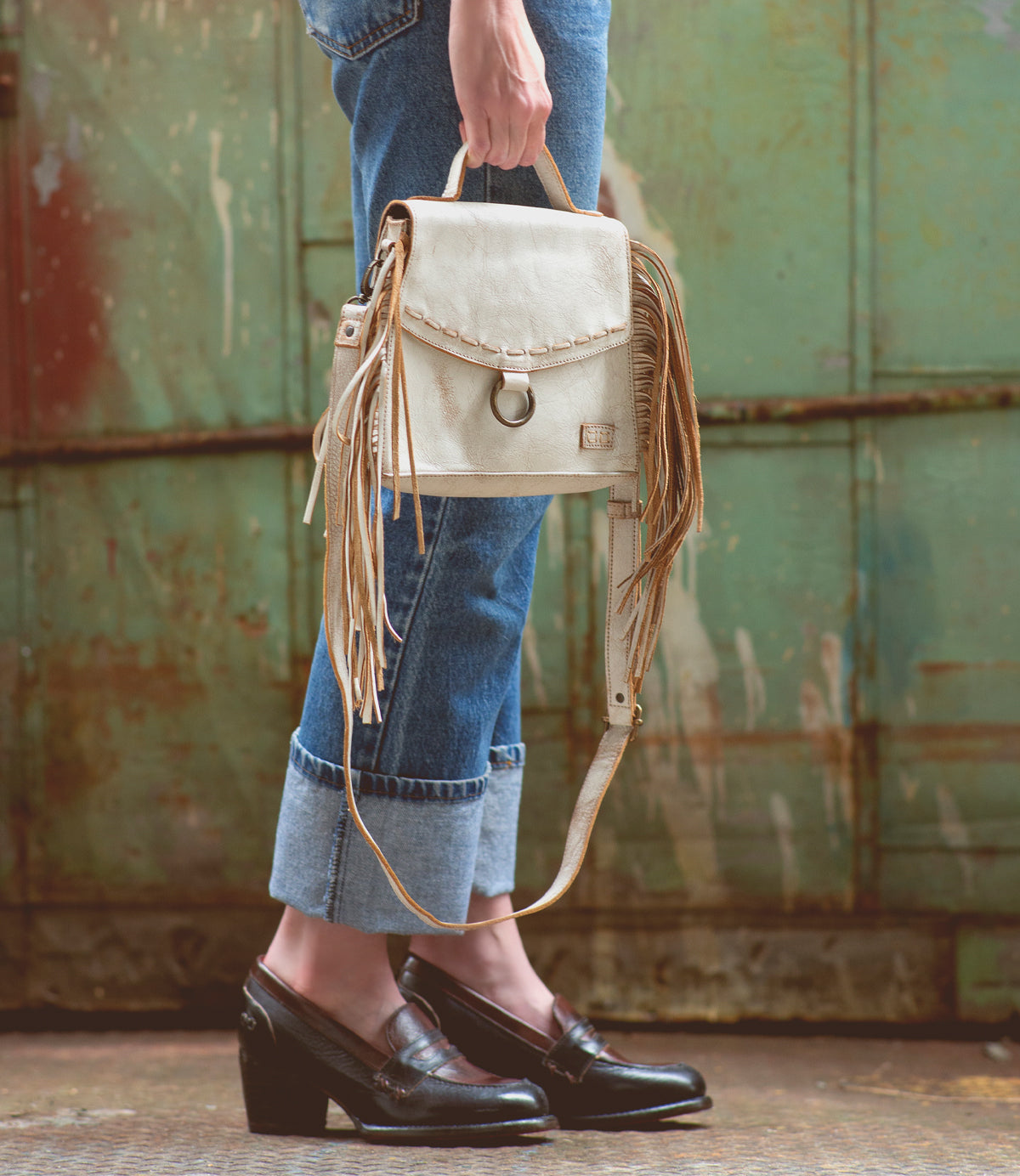 A person wearing cuffed jeans and black loafers holds the Bed|Stü Hidden beige fringed handbag with a structured silhouette in front of a weathered, green metal background.