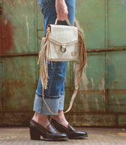 A person wearing cuffed jeans and black loafers holds the Bed|Stü Hidden beige fringed handbag with a structured silhouette in front of a weathered, green metal background.