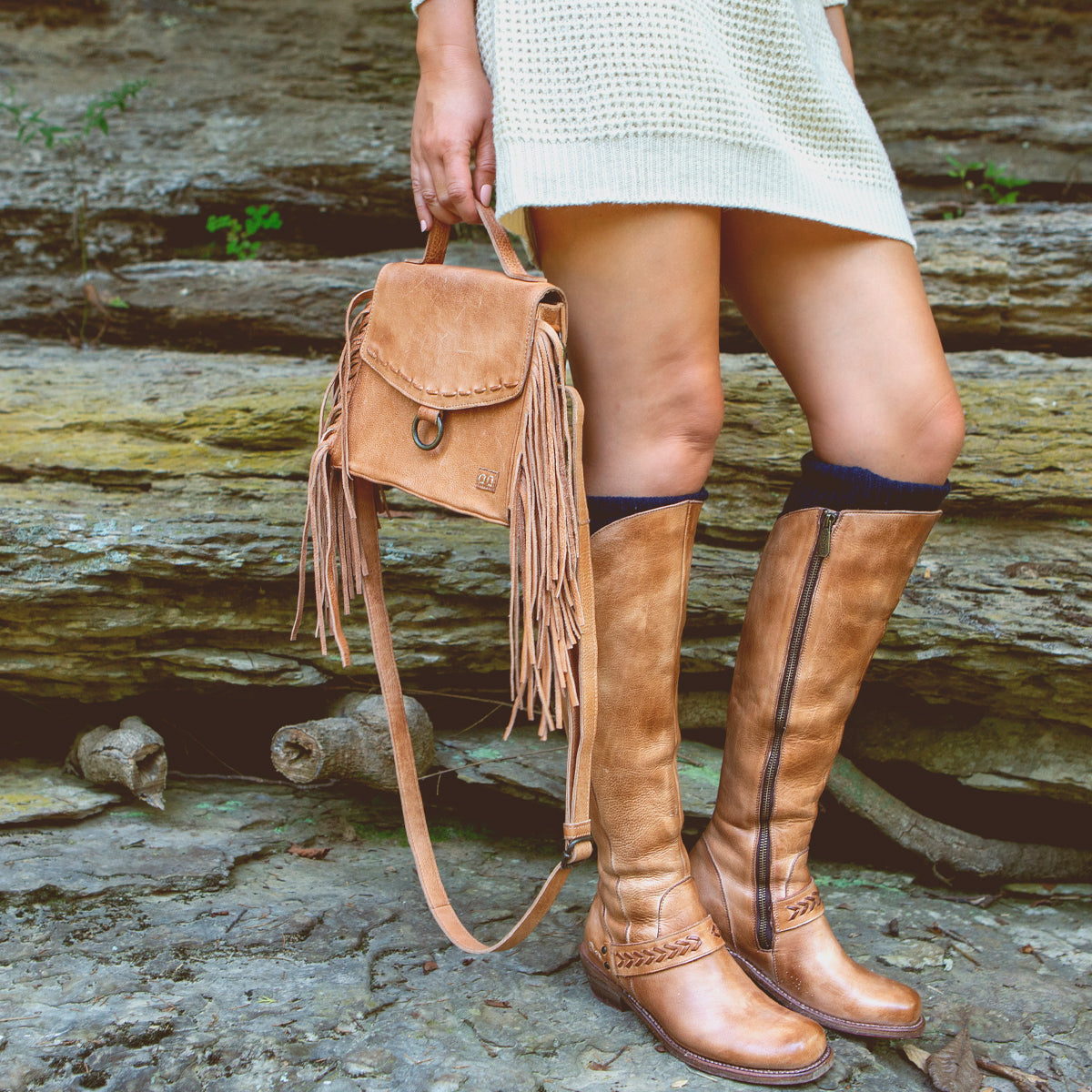A person wearing a cream sweater dress stands on rocky ground outdoors, holding the Bed|Stü Hidden backpack—a tan fringe bag with a soft leather finish—and wears matching tan knee-high boots.