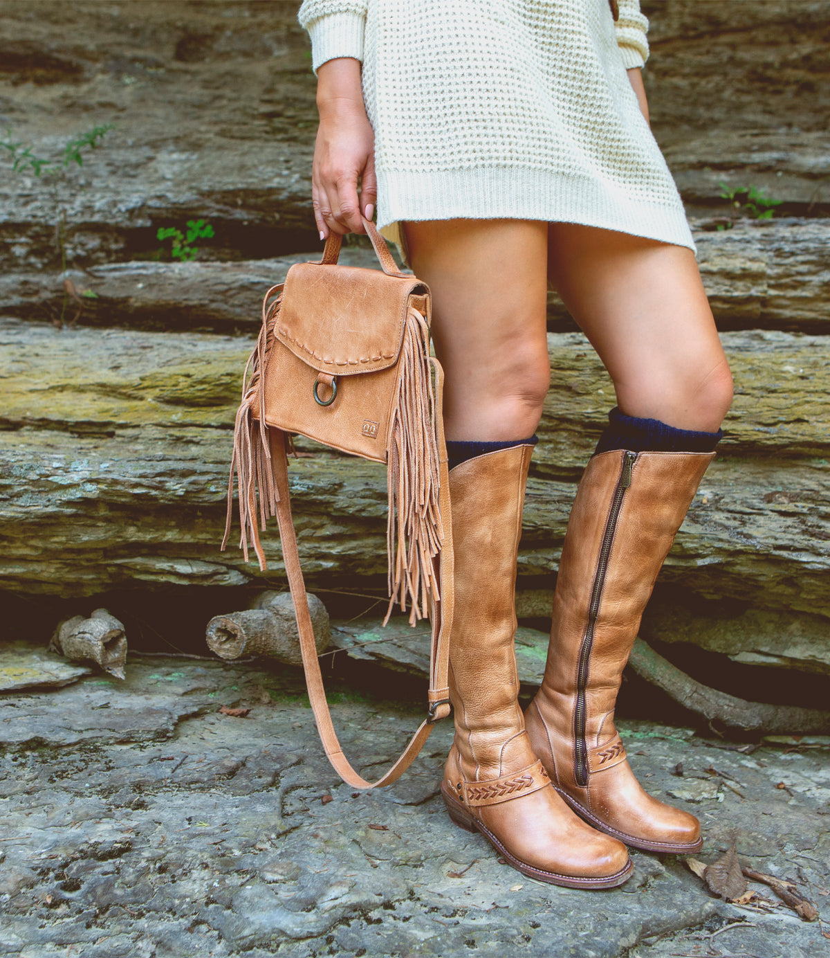A person wearing a cream sweater dress stands on rocky ground outdoors, holding the Bed|Stü Hidden backpack—a tan fringe bag with a soft leather finish—and wears matching tan knee-high boots.