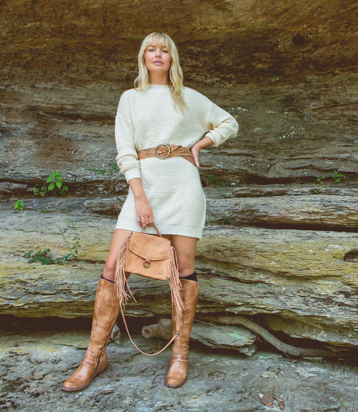 A woman stands in front of a rocky background wearing a cream-colored dress, brown belt, knee-high brown boots, and carrying the Bed|Stü Hidden brown fringe bag with a crossbody strap.