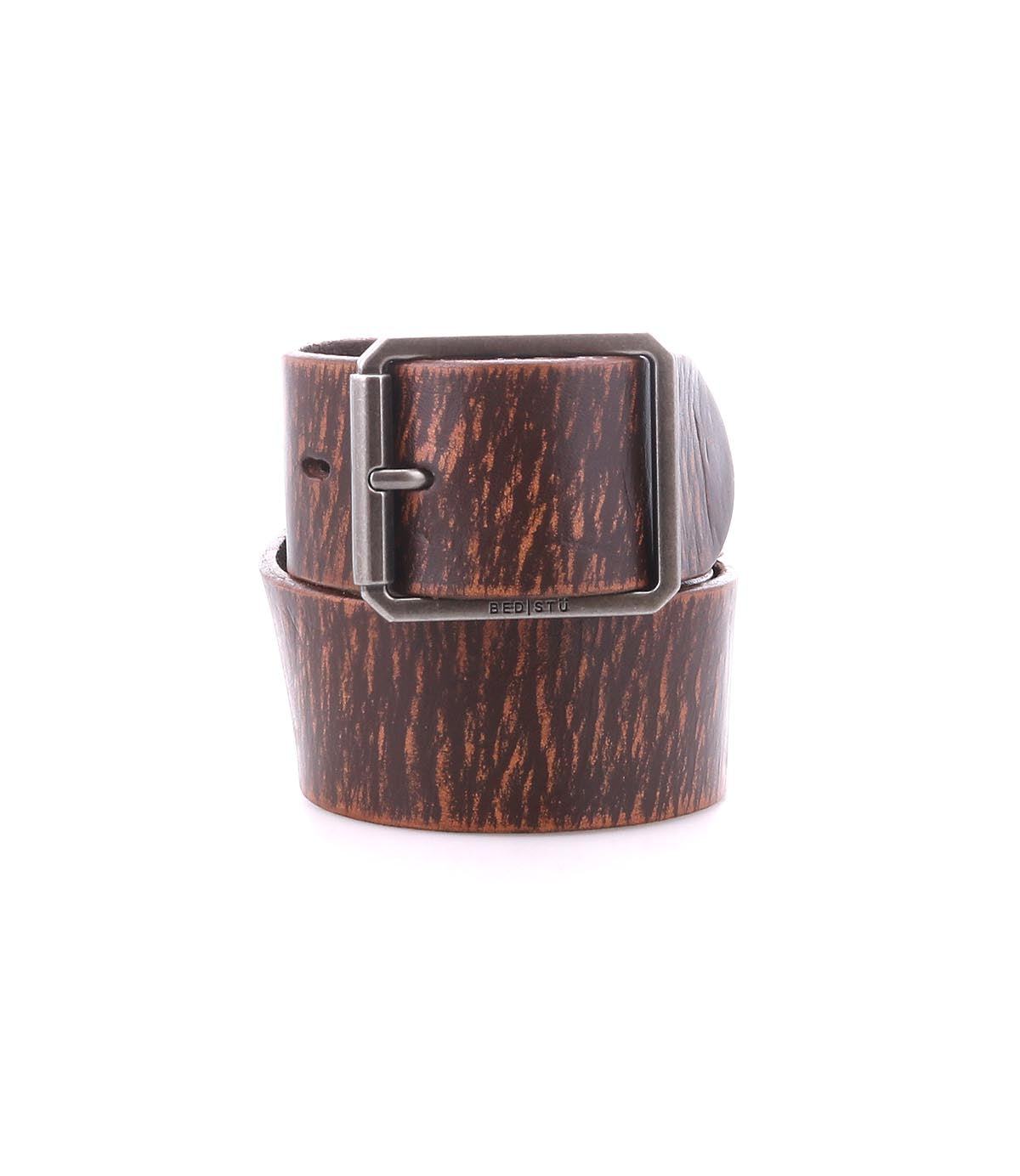 Introducing the Bed|Stü Hobo—a dark brown leather belt with a textured woodgrain pattern, crafted from a durable leather strap and finished with a sturdy metal buckle, rolled up on a white background—perfect for elevating your everyday style.