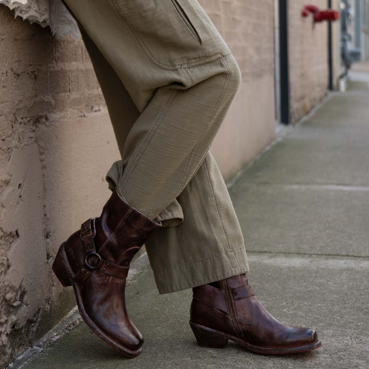 Wearing the Bed|Stü Homage rich leather boots featuring a harness strap, this person in olive cargo pants leans casually against a brick wall on the sidewalk.