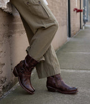Wearing the Bed|Stü Homage rich leather boots featuring a harness strap, this person in olive cargo pants leans casually against a brick wall on the sidewalk.