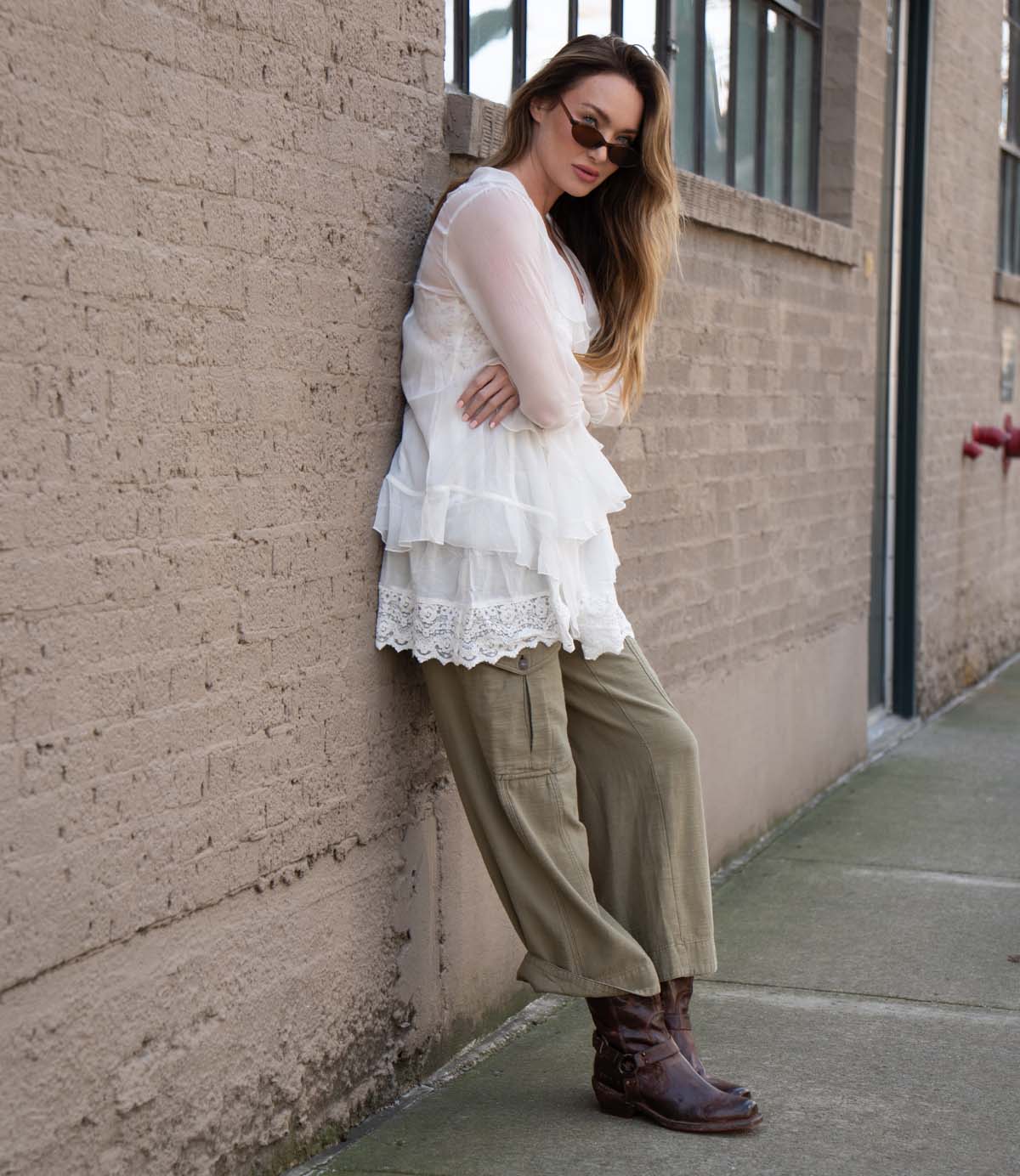 A woman leans against a brick wall on a city sidewalk, wearing a white layered blouse, olive green pants, sunglasses, and finishing her look with Bed|Stü’s Homage rich leather boots.