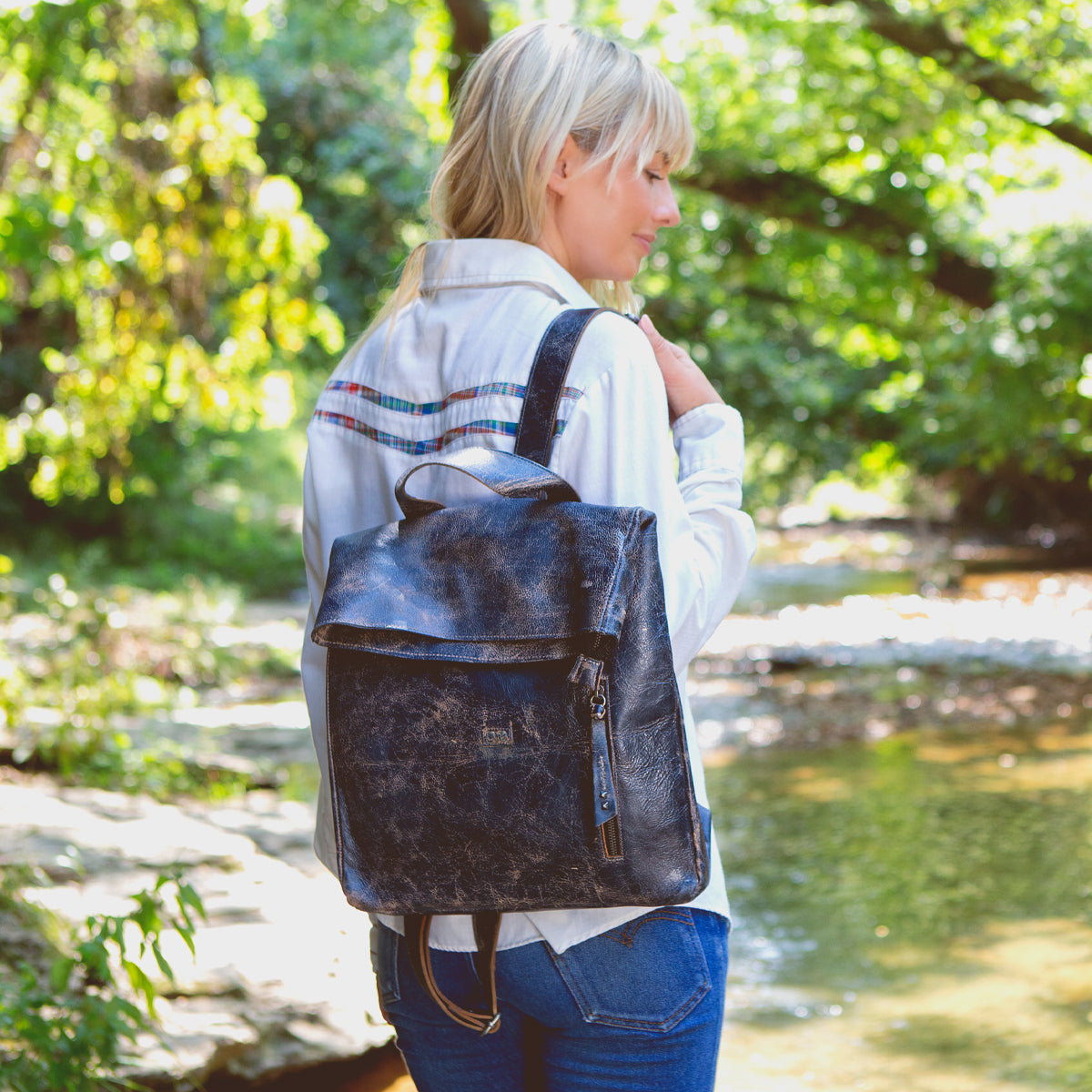 A person with blonde hair wearing a white shirt and jeans stands near a creek, carrying the Bed|Stü Howie black leather laptop backpack with adjustable straps.