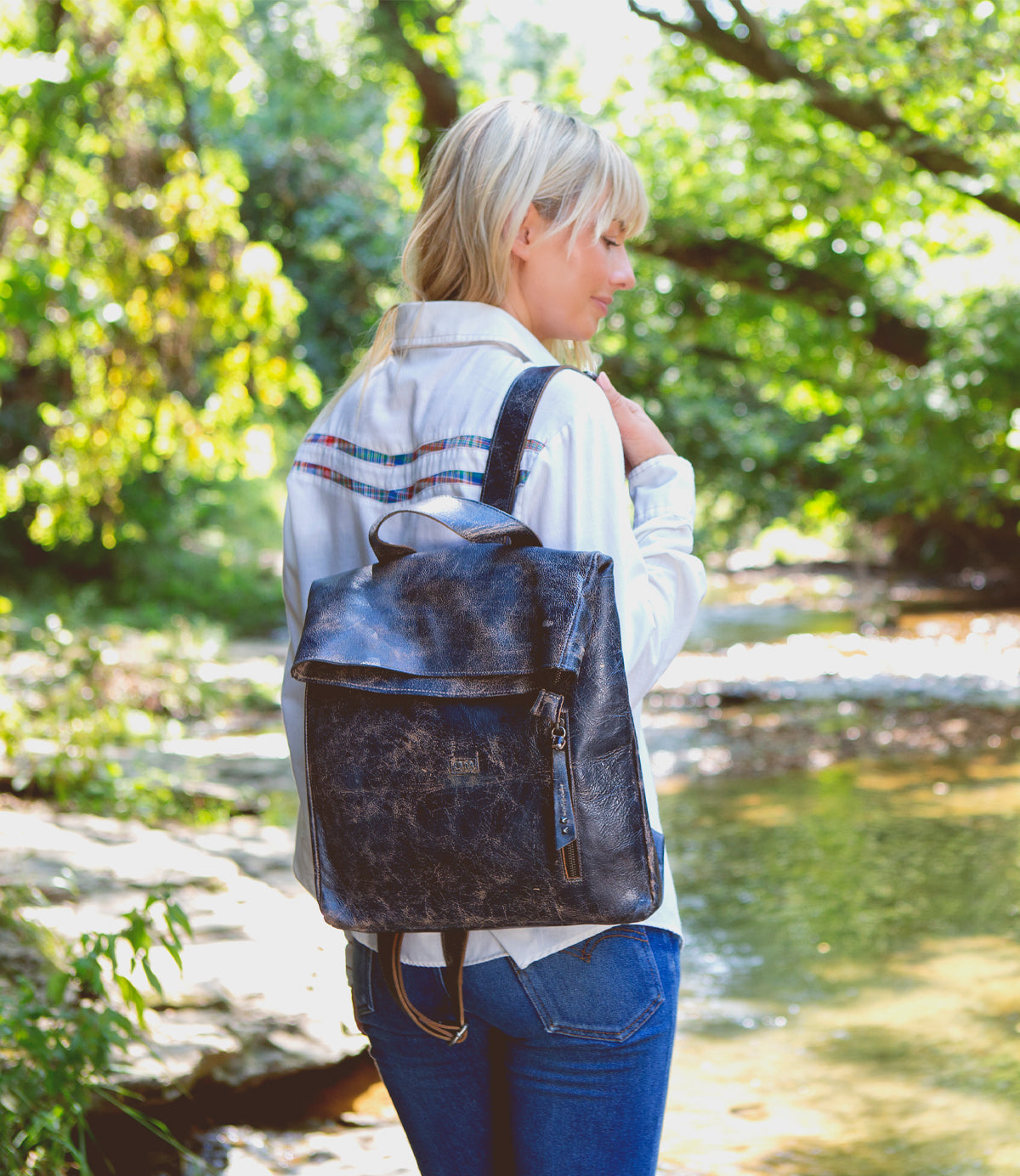 A person with blonde hair wearing a white shirt and jeans stands near a creek, carrying the Bed|Stü Howie black leather laptop backpack with adjustable straps.