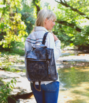 A person with blonde hair wearing a white shirt and jeans stands near a creek, carrying the Bed|Stü Howie black leather laptop backpack with adjustable straps.