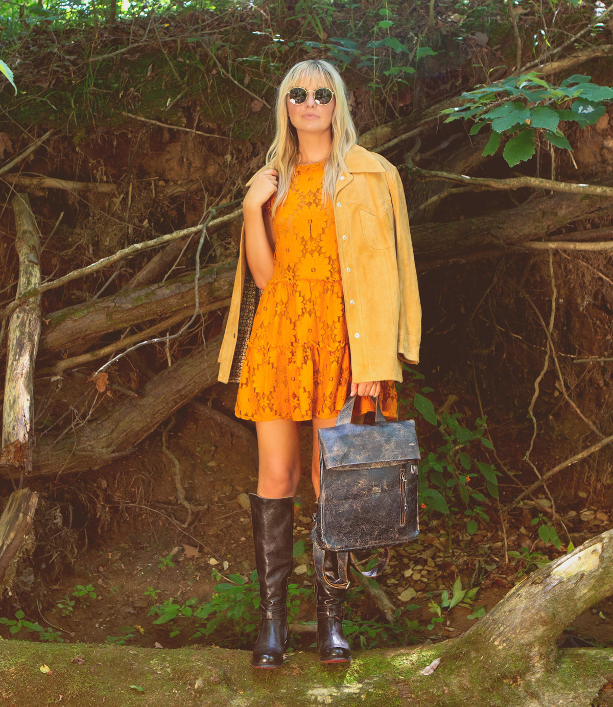 A woman in a yellow dress, tan jacket, and black boots stands outdoors among trees and roots, wearing sunglasses and holding the Bed|Stü Howie—a sleek leather backpack with adjustable straps.