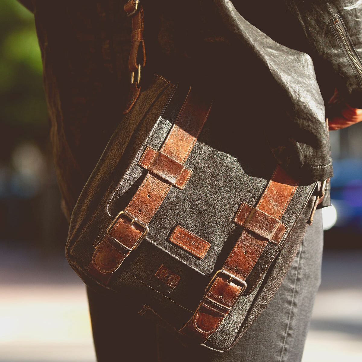 Wearing a black leather jacket and jeans, this person carries the Bed|Stü Hue messenger bag—a sleek black design featuring brown leather straps, metal buckles, and an adjustable crossbody strap for effortless style from work to weekend.