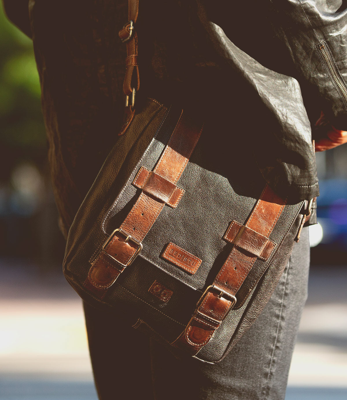 Wearing a black leather jacket and jeans, this person carries the Bed|Stü Hue messenger bag—a sleek black design featuring brown leather straps, metal buckles, and an adjustable crossbody strap for effortless style from work to weekend.