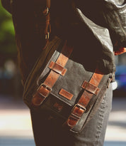 Wearing a black leather jacket and jeans, this person carries the Bed|Stü Hue messenger bag—a sleek black design featuring brown leather straps, metal buckles, and an adjustable crossbody strap for effortless style from work to weekend.