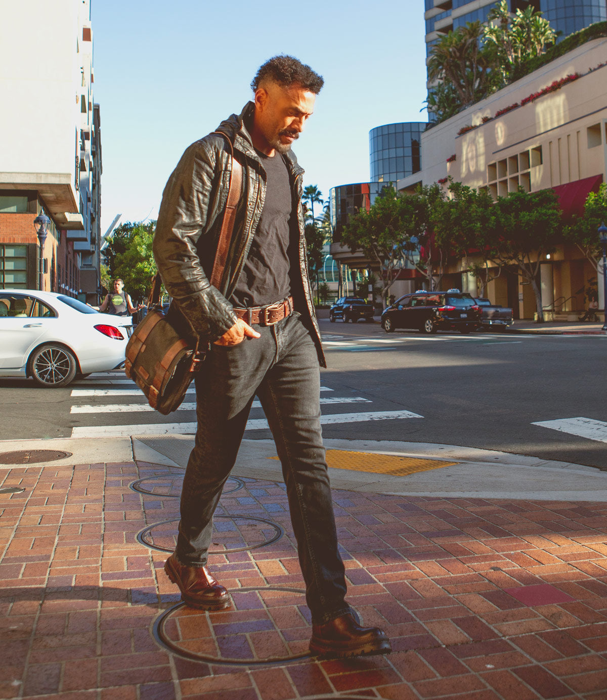A man wearing dark jeans, a black shirt, a leather jacket, and boots walks across a city crosswalk with the Bed|Stü Hue messenger bag—featuring an adjustable crossbody strap—slung over his shoulder. This versatile accessory transitions effortlessly from work to weekend. Daylight, buildings, and cars set the urban scene in the background.