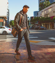 A man wearing dark jeans, a black shirt, a leather jacket, and boots walks across a city crosswalk with the Bed|Stü Hue messenger bag—featuring an adjustable crossbody strap—slung over his shoulder. This versatile accessory transitions effortlessly from work to weekend. Daylight, buildings, and cars set the urban scene in the background.