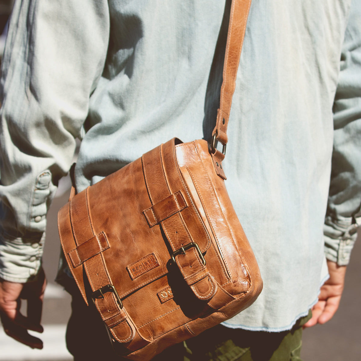 A person wearing a light denim shirt and green pants, shown from behind, carries the Bed|Stü Hue bag—a brown leather messenger with buckle straps and an adjustable crossbody strap—over their shoulder, making it perfect for seamlessly transitioning from work to weekend.