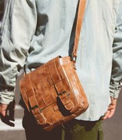 A person wearing a light denim shirt and green pants, shown from behind, carries the Bed|Stü Hue bag—a brown leather messenger with buckle straps and an adjustable crossbody strap—over their shoulder, making it perfect for seamlessly transitioning from work to weekend.