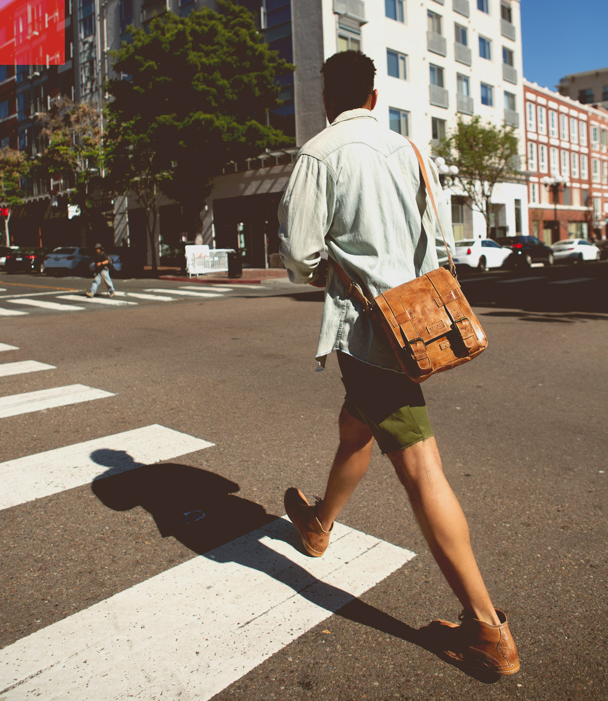 A person wearing a light shirt, shorts, and brown shoes carries the Bed|Stü Hue messenger bag—with its adjustable crossbody strap—as they cross a city street on a sunny day, effortlessly blending work-to-weekend style.